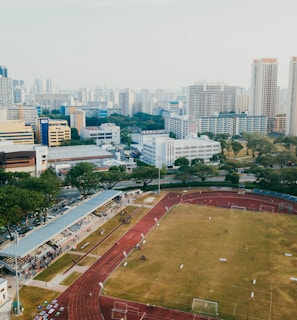 An aerial view of a sports complex with a running track surrounding a grassy field, set against the background of a cityscape. The city's skyline features a mix of tall residential buildings and shorter commercial structures. There are trees and green areas surrounding the sports facility.