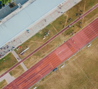 An aerial view of a sports complex featuring a running track and a grassy field. The tracks are red and situated next to a large building. There are several people scattered across the area, some on the track and others near the building. The surroundings include trees and geometric pathways, adding structure to the scene.
