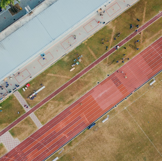 An aerial view of a sports complex featuring a running track and a grassy field. The tracks are red and situated next to a large building. There are several people scattered across the area, some on the track and others near the building. The surroundings include trees and geometric pathways, adding structure to the scene.