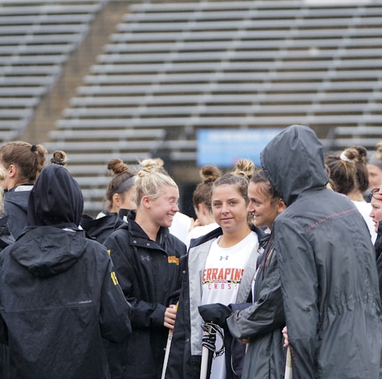 A group of people, likely athletes, are gathered together on a sports field. They are wearing black jackets with some individuals dressed in white sports uniforms underneath. A few people have their hair tied up in buns, and most are engaged in conversation. The background shows tiered stadium seats, suggesting the setting is an outdoor sports venue.