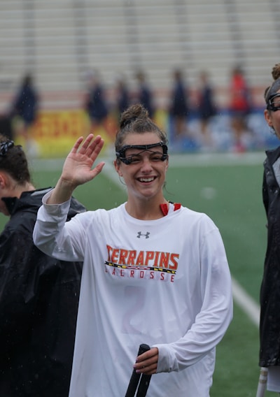 Several athletes in sports attire are on an outdoor sports field, interacting and smiling. One individual in the foreground waves with a friendly expression, while another stands beside her, also smiling. They wear protective goggles and gear associated with lacrosse. The background features indistinct figures in dark clothing against a backdrop of stadium seating.