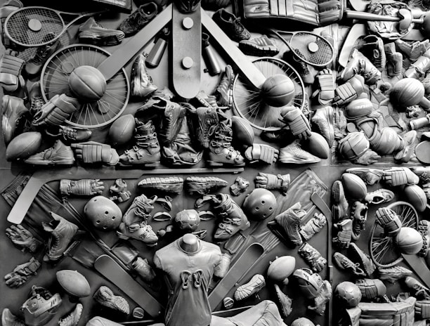 A complex arrangement of various sports equipment and attire, including tennis rackets, soccer balls, basketballs, helmets, shoes, gloves, and a mannequin torso wearing a T-shirt. The items are layered and artistically displayed in a monochrome color scheme, creating an intricate texture and visual pattern.