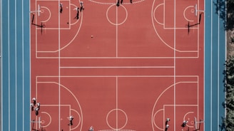 An aerial view of a large, vibrant sports complex featuring a red basketball court with multiple hoops and a surrounding blue running track. Several people are scattered across the area, engaged in various activities.
