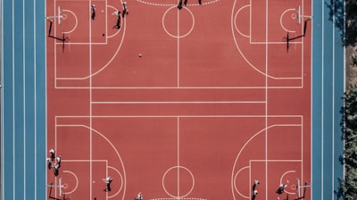 An aerial view of a large, vibrant sports complex featuring a red basketball court with multiple hoops and a surrounding blue running track. Several people are scattered across the area, engaged in various activities.