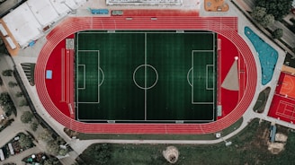 An aerial view of a large sports complex featuring a central green football field surrounded by a red running track. Adjacent to the track, there is a parking area, buildings, and pathways. Nearby, there are additional sports courts in different colors, including a blue swimming pool and orange basketball courts.