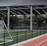 An outdoor sports facility featuring a running track and a soccer goal. The track is red, surrounded by a black metal fence, and the field is covered with artificial green turf. In the background, a basketball court is visible under a covered roof with metal supports. The sky is overcast, adding a moody atmosphere.