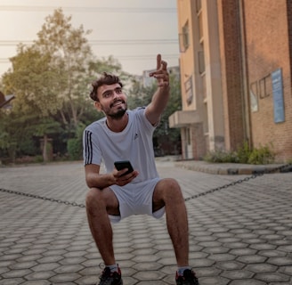 A man in a white sports outfit sits on a low chain while holding a smartphone. He is smiling and gesturing upward with his right hand. The background includes a brick building to the right, greenery, and a paved area with trees and power lines visible in the distance.