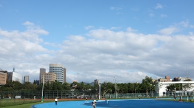 A bright outdoor sports complex features a blue running track, surrounded by green grass and trees. Several people are visible, engaged in activities on the track. In the background, urban high-rise buildings rise against a partly cloudy sky.