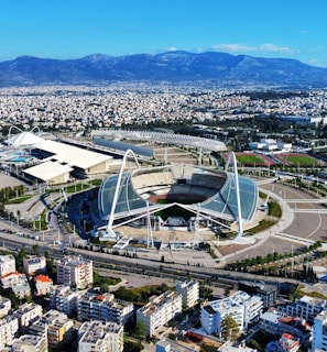 A large, modern sports complex is situated within an expansive urban landscape. The main stadium features prominent architectural structures and an open roof design. Surrounding the stadium are various sports facilities, roads, and greenery. Residential and commercial buildings extend into the distance, framed by mountain ranges under a clear blue sky.
